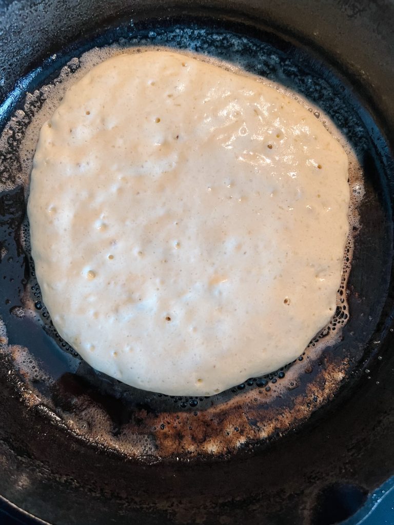 pancake on skillet. The batter is bubbling up indicating it's ready to flip over to the other side.
