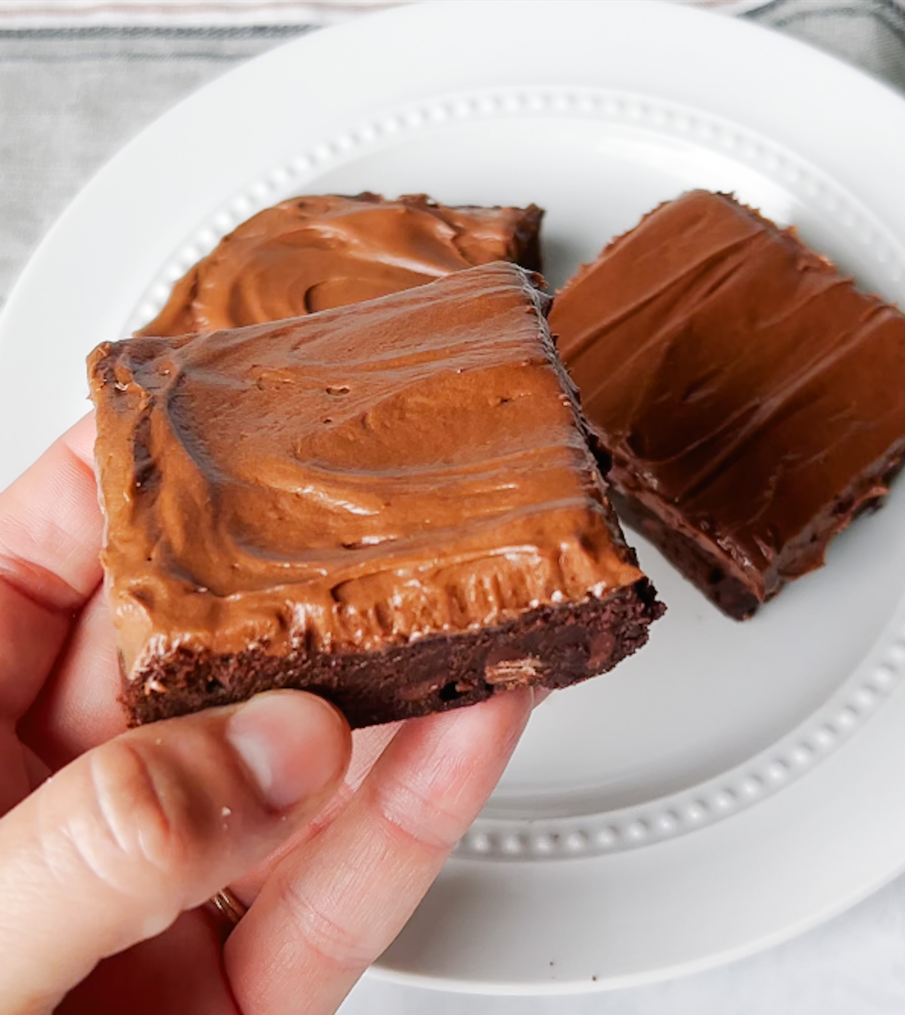 Holding one frosted brownie with two brownies sitting on a plate in the background.