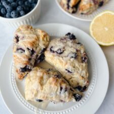 Three buttermilk lemon blueberry scones on a plate with a lemon and a bowl of blueberries in the background.