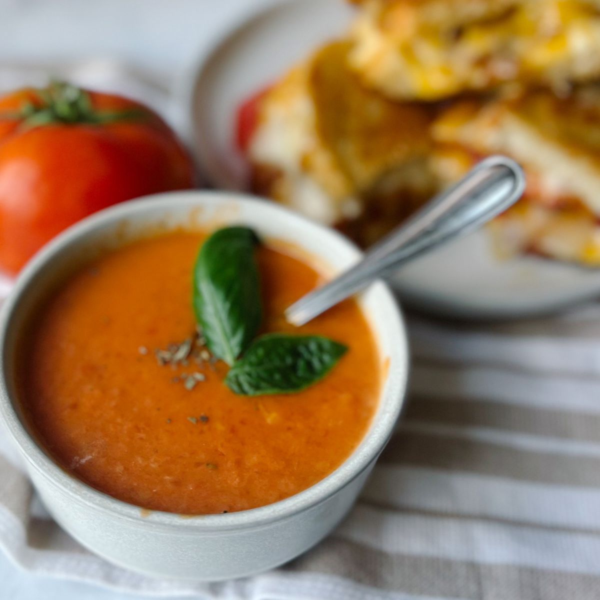 Tomato soup topped with fresh basil leaves and a plate of grilled cheese behind it.