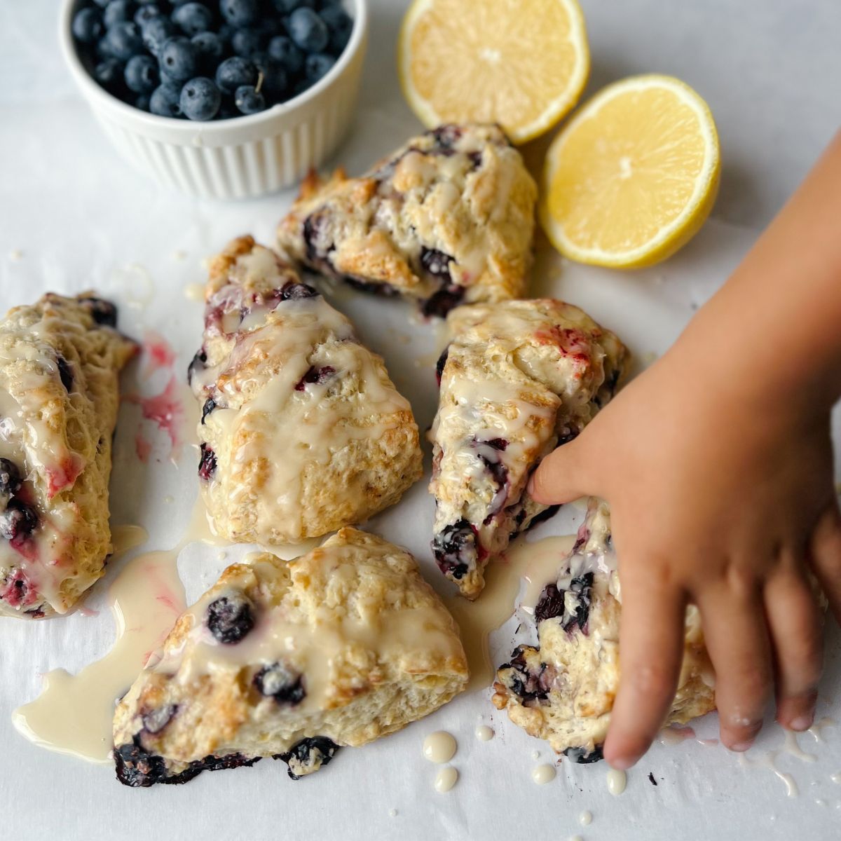Buttermilk lemon blueberry scones sitting on parchment paper with a child's hand reaching toward one with lemons and blueberries in the background.