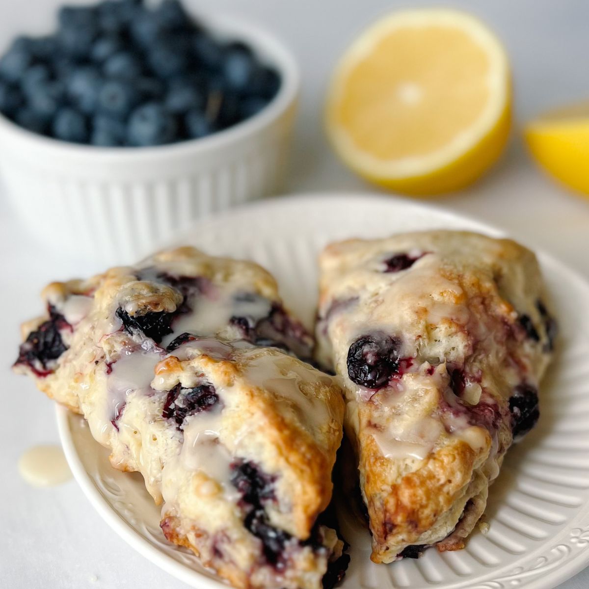 two lemon blueberry scones on a white plate with two half lemons and a bowl of blueberries in the background.