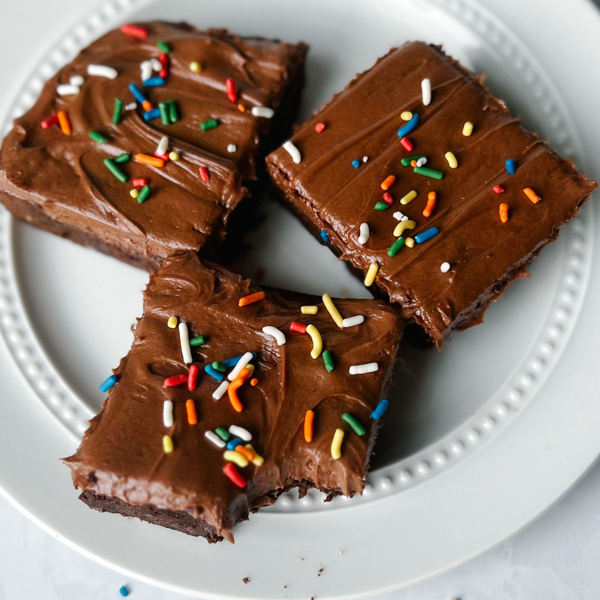 Plate of three brownies with chocolate frosting and sprinkles on top. One brownie has a bite taken out of it.