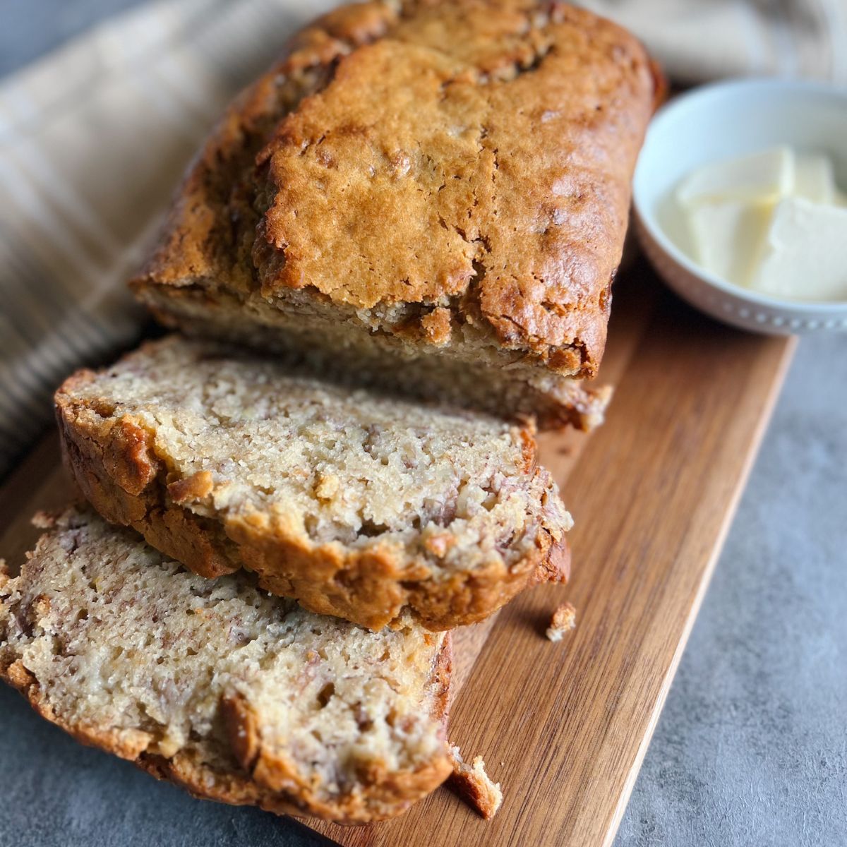 Tahini banana bread loaf with two slices already cut. It sits on a wooden cutting board with a dish of butter pads in the background.