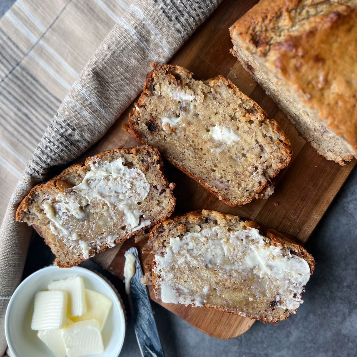 Three slices of tahini banana bread with butter spread on them. They are sitting on top of a wooden cutting board with a dish of butter pads and the loaf next to them.