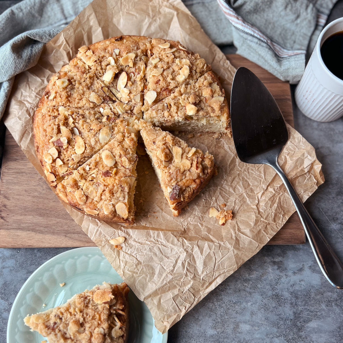 Almond coffee cake on parchment paper and a cutting board with a slice of coffee cake on a plate near it and a cup of coffee on the top right corner.