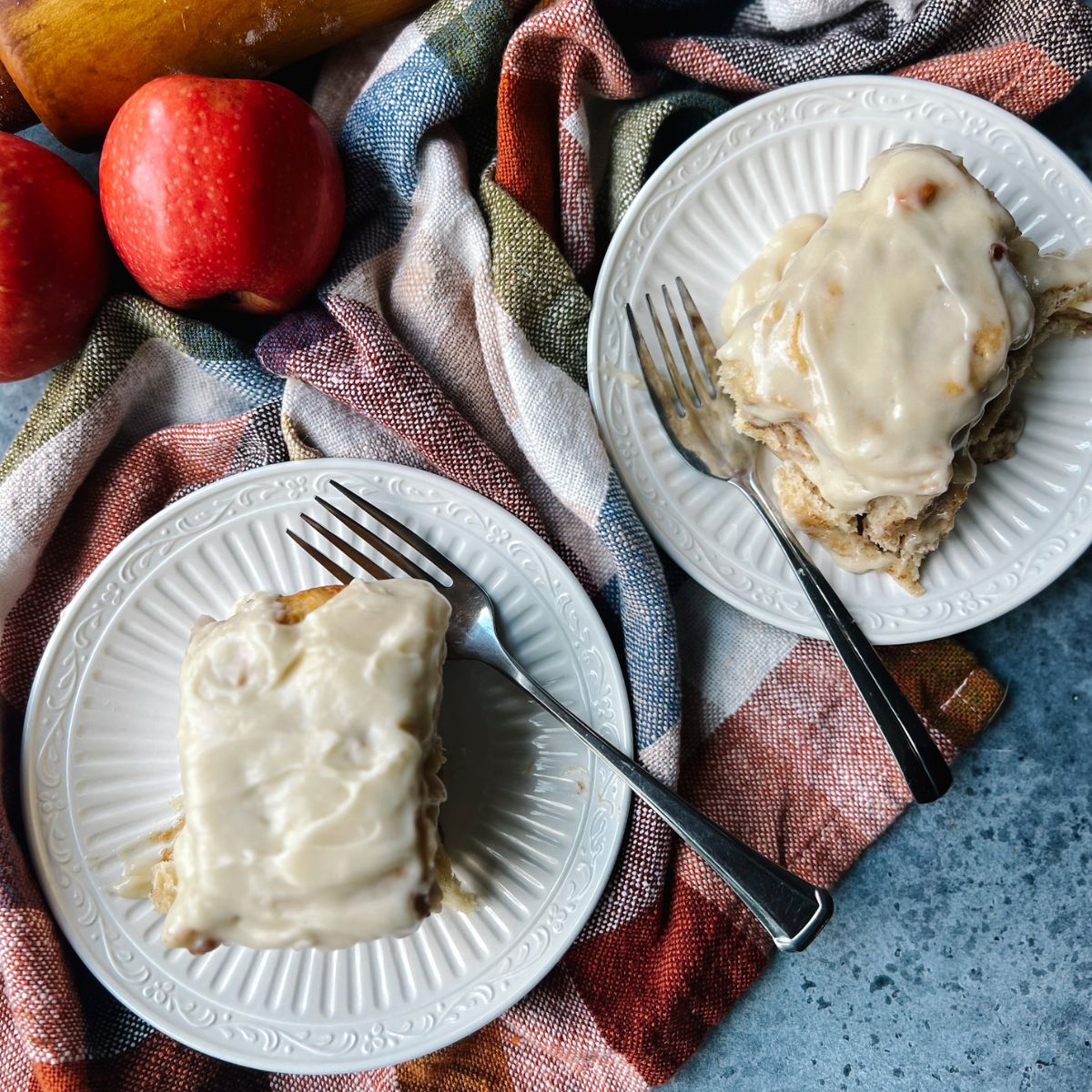 Two cinnamon rolls with caramel cream cheese frosting on plates. There is a plaid tea towel and two apples laid next to them.