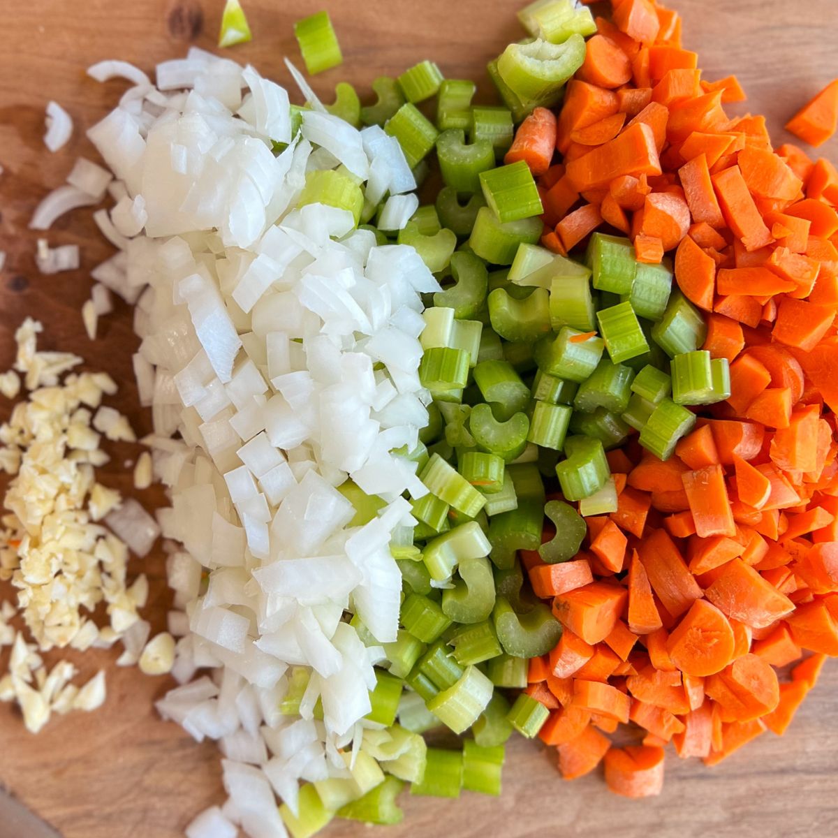 Chopped onion, celery, carrots, and garlic on a wooden cutting board.