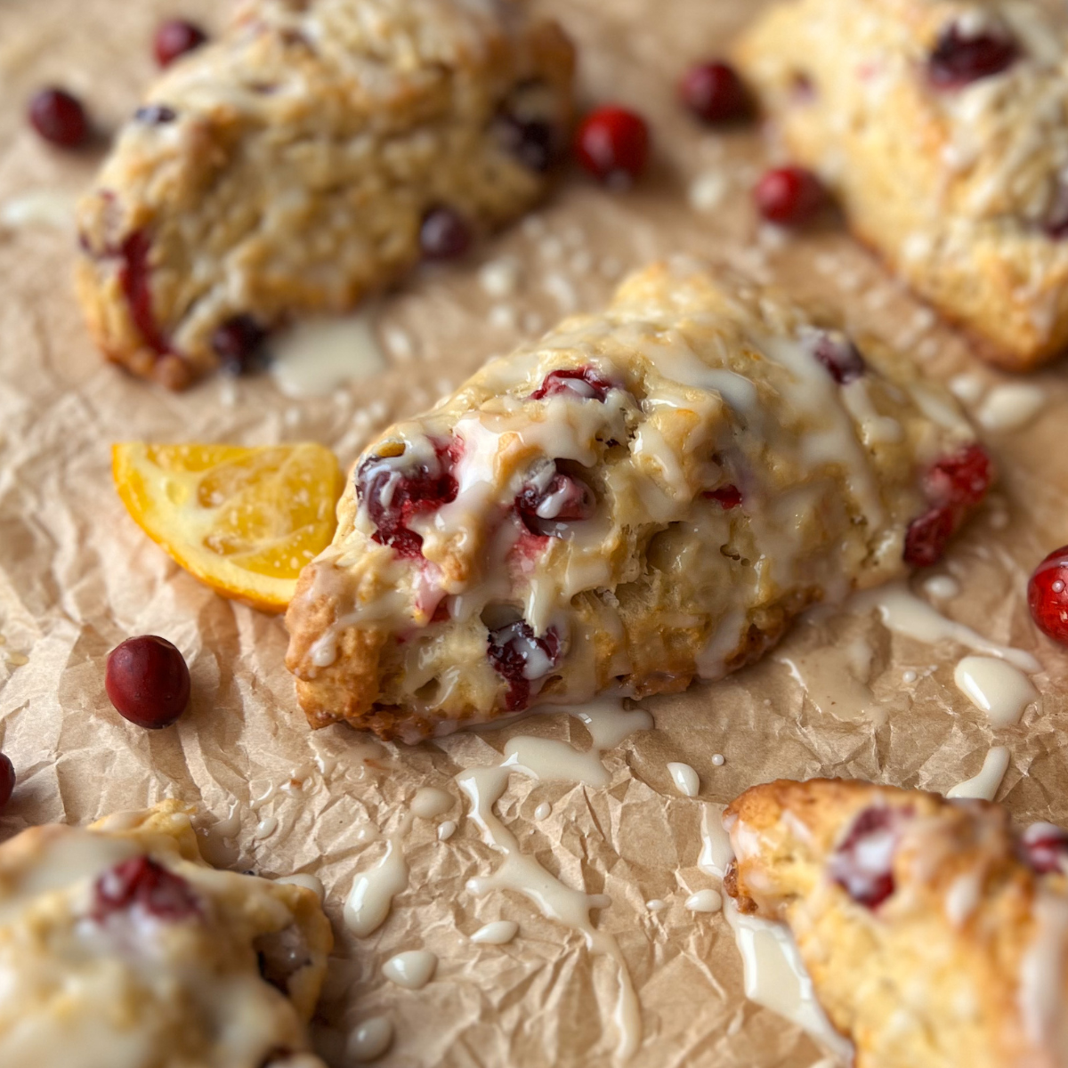 Close up shot of the orange cranberry scones topped with orange glaze. Several of them sit on parchment paper with cranberries and oranges adorning the paper.