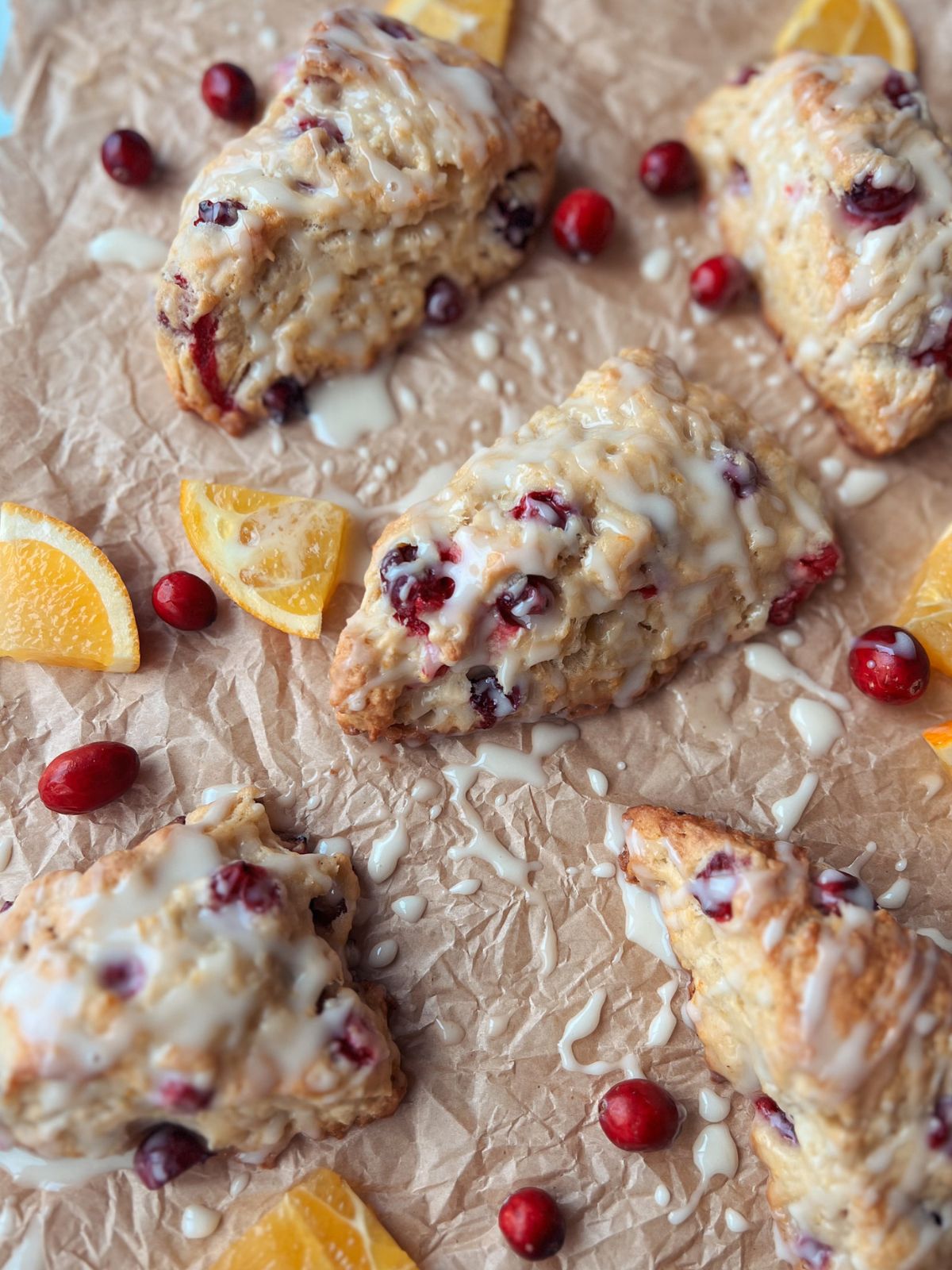Five cranberry orange scones with orange glaze on parchment paper surrounded by cranberries and orange slices.