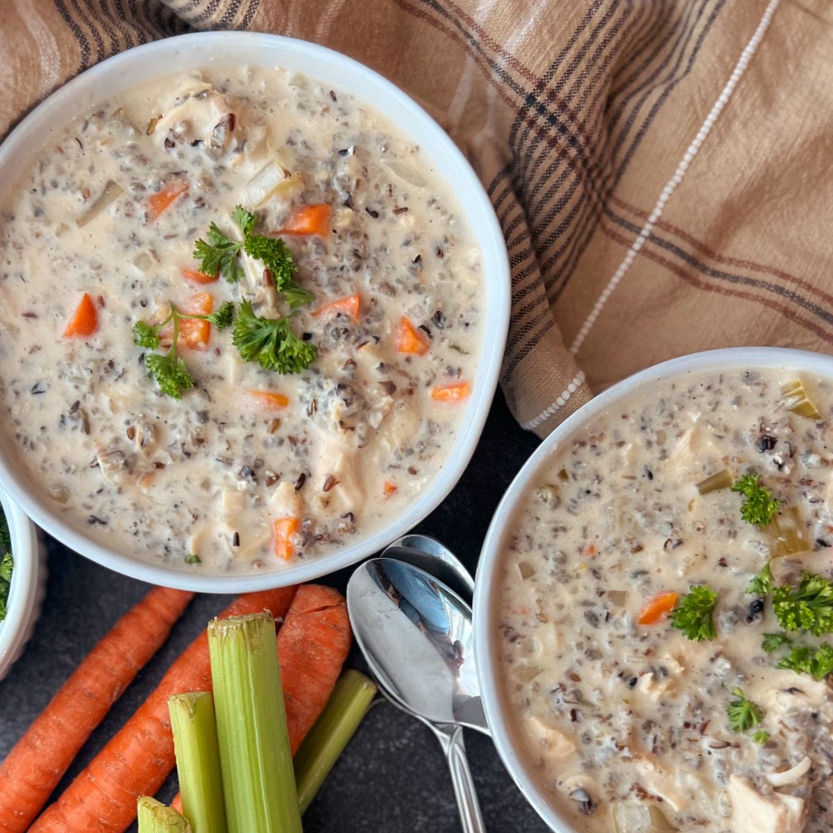 Two bowls of creamy chicken wild rice soup with carrots and celery and a towel near them.