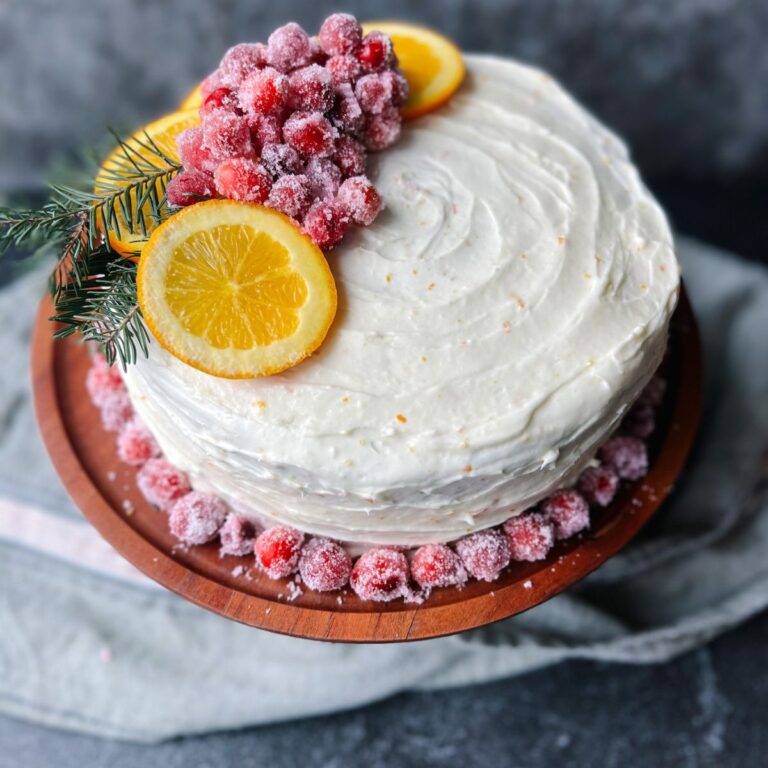 Cranberry orange cake on a wooden cake stand with a kitchen towel in the background. The cake is decorated with sugared cranberries and orange slices.