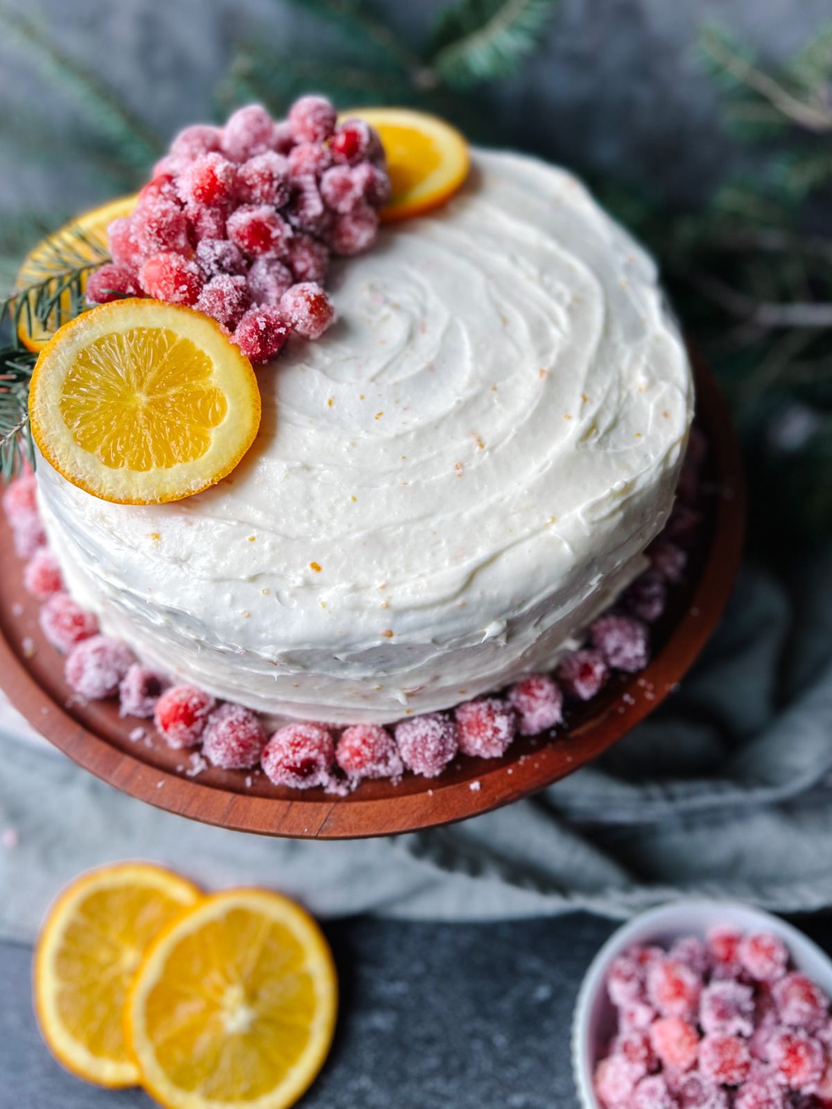 Cranberry orange cake on a wooden cake stand with spruce in the background and orange slices and sugared cranberries in the foreground.
