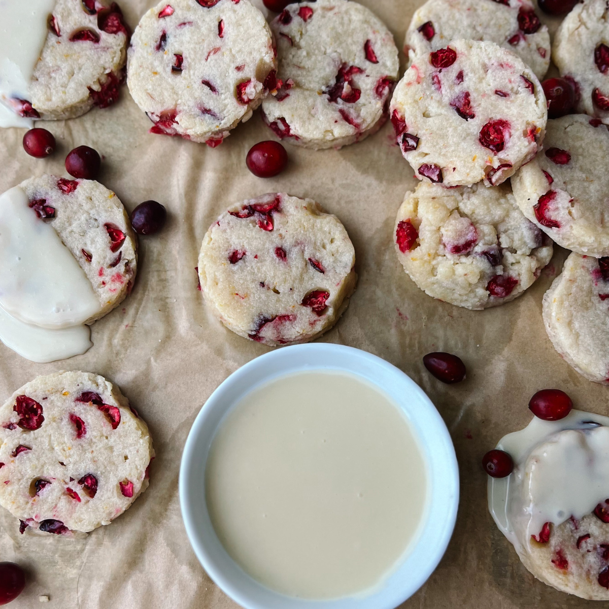 A bowl of melted white chocolate on parchment paper surrounded by cranberry orange shortbread cookies. 