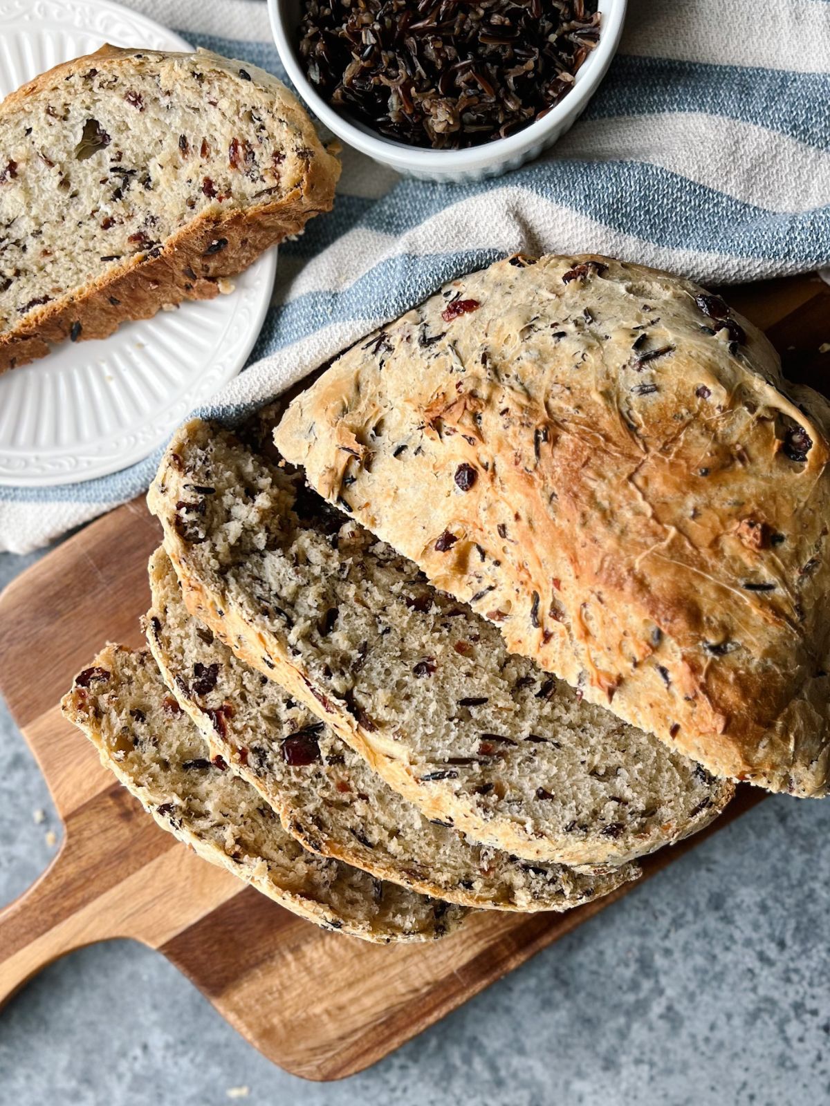 Loaf of cranberry wild rice bread on a wooden cutting board with a slice of bread on a plate next to it.