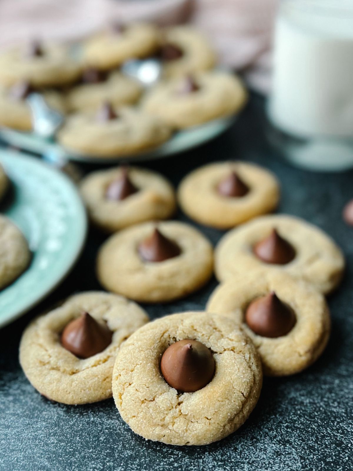 Peanut butter blossoms on a counter with a plate of more cookies and a glass of milk in the background.