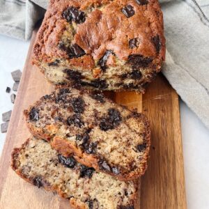 Chocolate chunk banana bread on a wooden cutting board with two slices cut out and on the side of it.