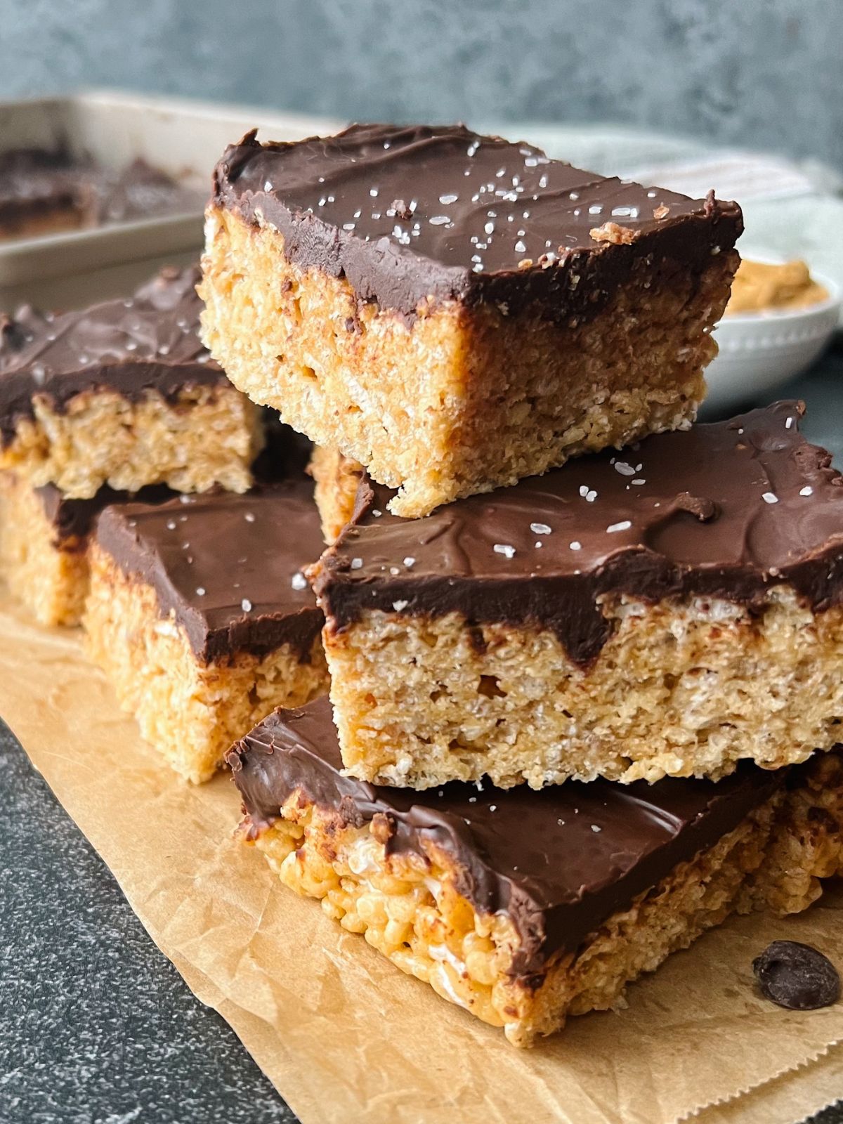 Several chocolate peanut butter krispie treats stacked on each other with a pan of the treats in the background.
