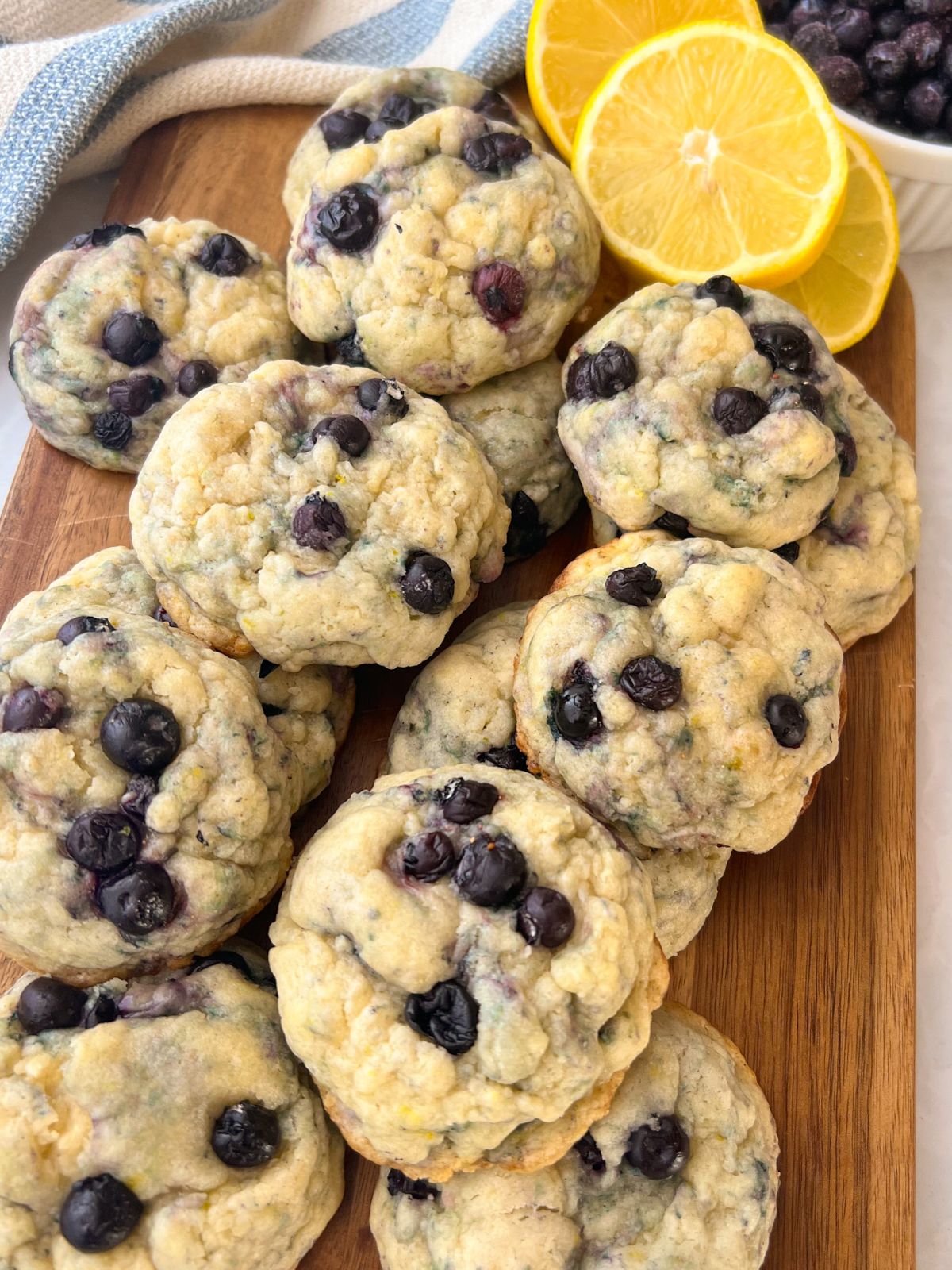 13 lemon blueberry cookies on a cutting board with a bowl of blueberries and cut lemons in the background.