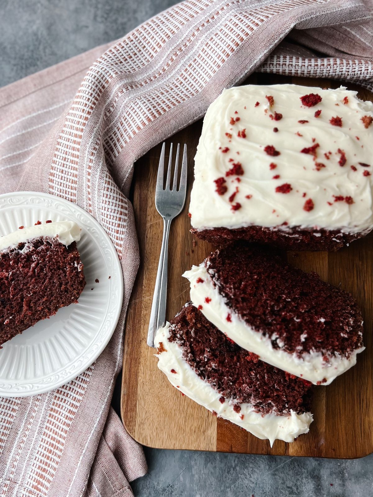 Red velvet loaf cake with three slices cut. Two of the slices sit with the loaf cake on a wooden cutting board. The third is on a plate next to it.