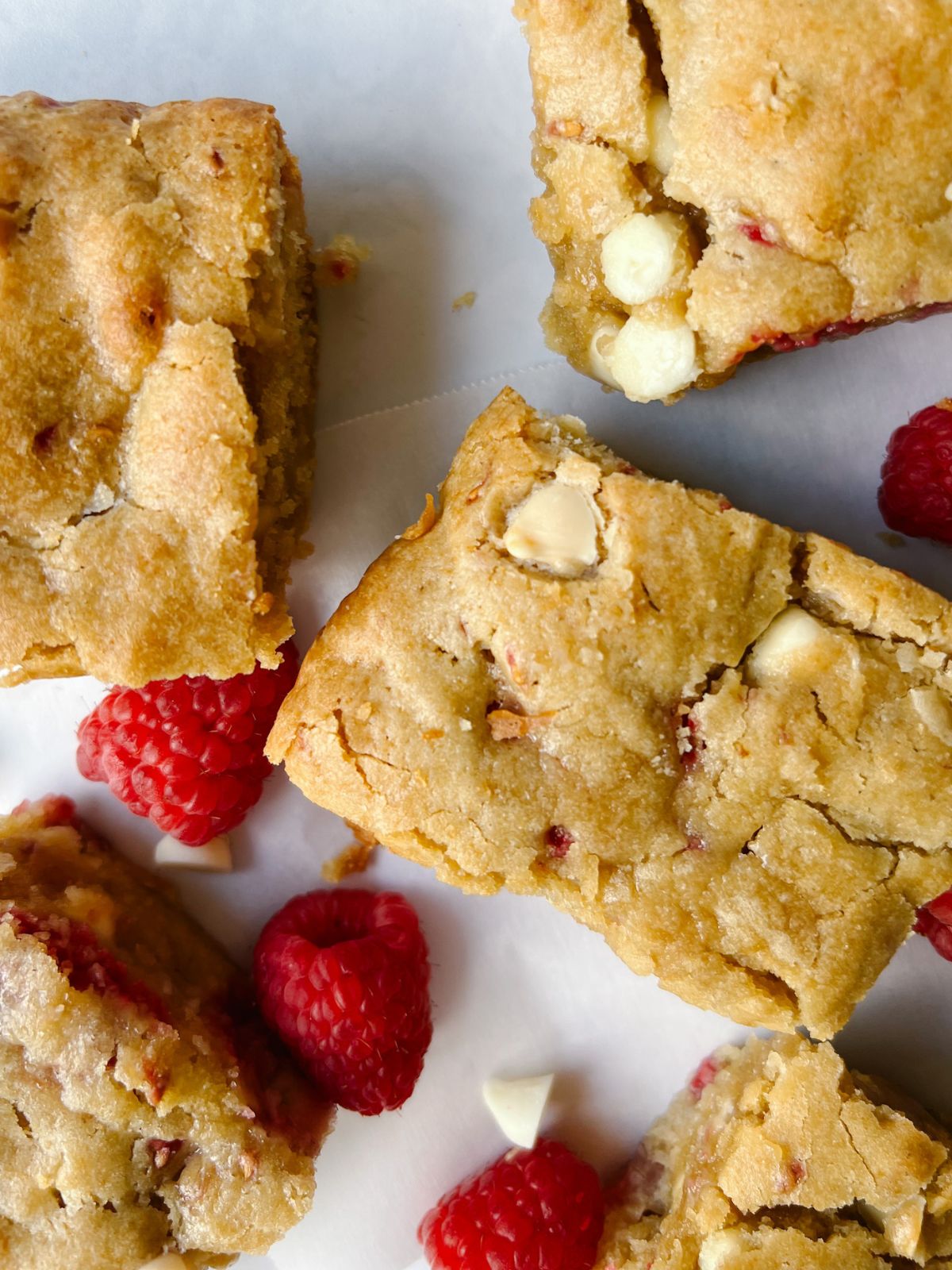 Several white chocolate raspberry blondies on parchment paper surrounded by white chocolate chips and raspberries.