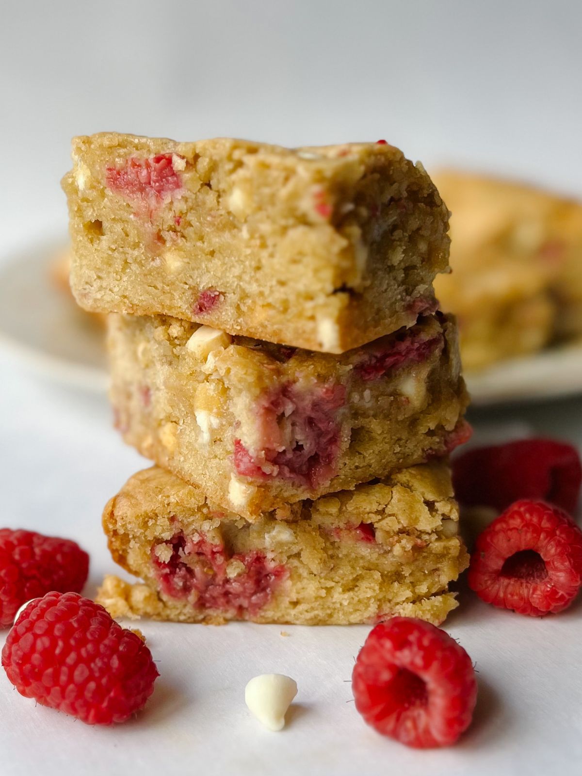 Three white chocolate raspberry blondies stacked on top of each other with a plate of more blondies in the background.