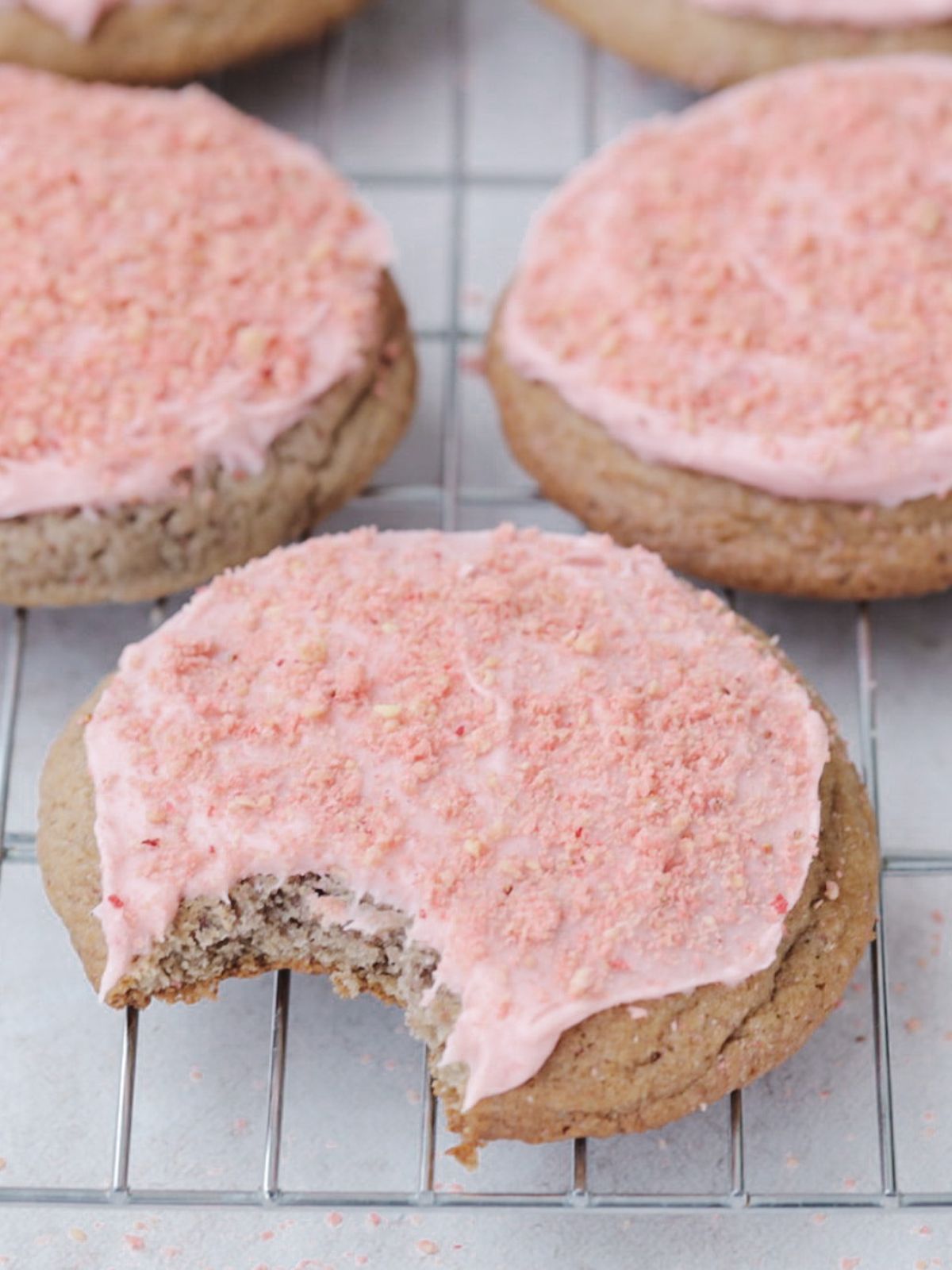 Three  strawberry crunch cookies with strawberry cream cheese frosting on a wire rack with the first cookie having had a bite taken out of it.