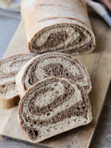 Three sourdough marble rye bread slices are shown on a wooden cutting board with the remainder of the sourdough rye bread in the background. You can see the closeup of the marbled swirl in the bread.