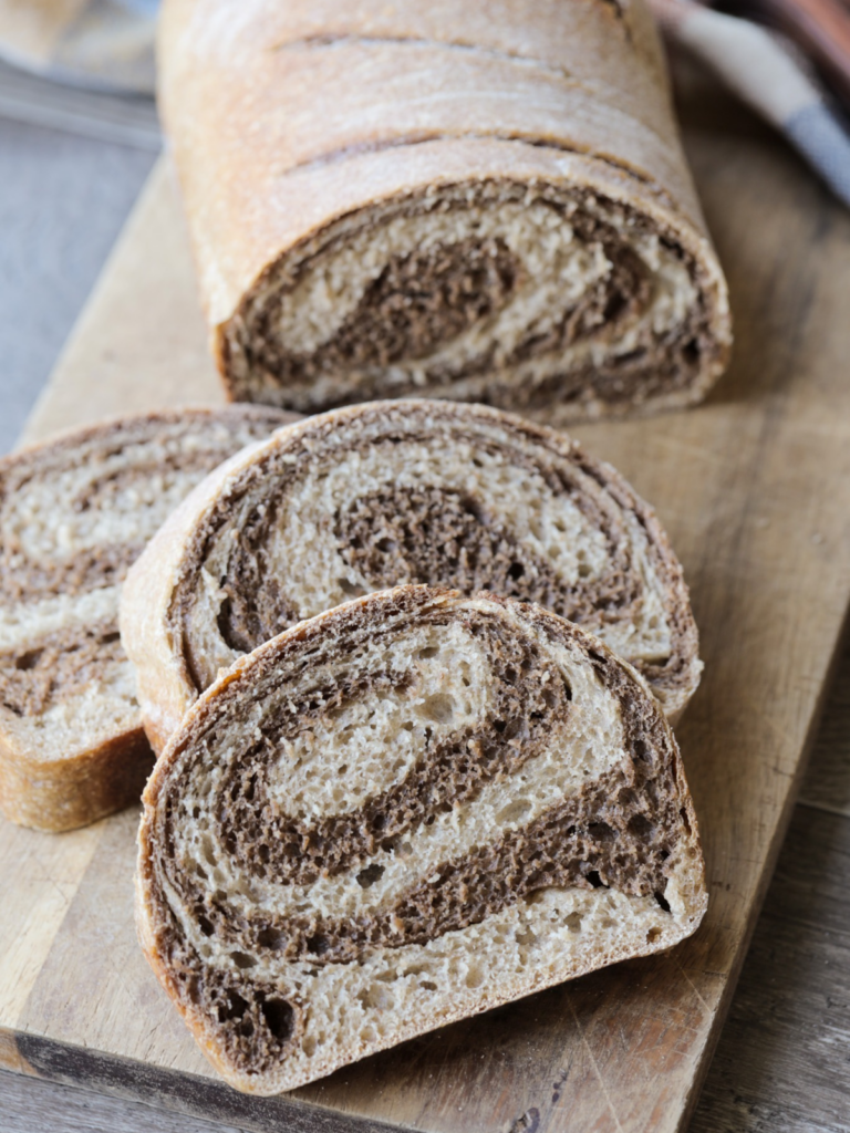 Three sourdough marble rye bread slices are shown on a wooden cutting board with the remainder of the sourdough rye bread in the background. You can see the closeup of the marbled swirl in the bread.