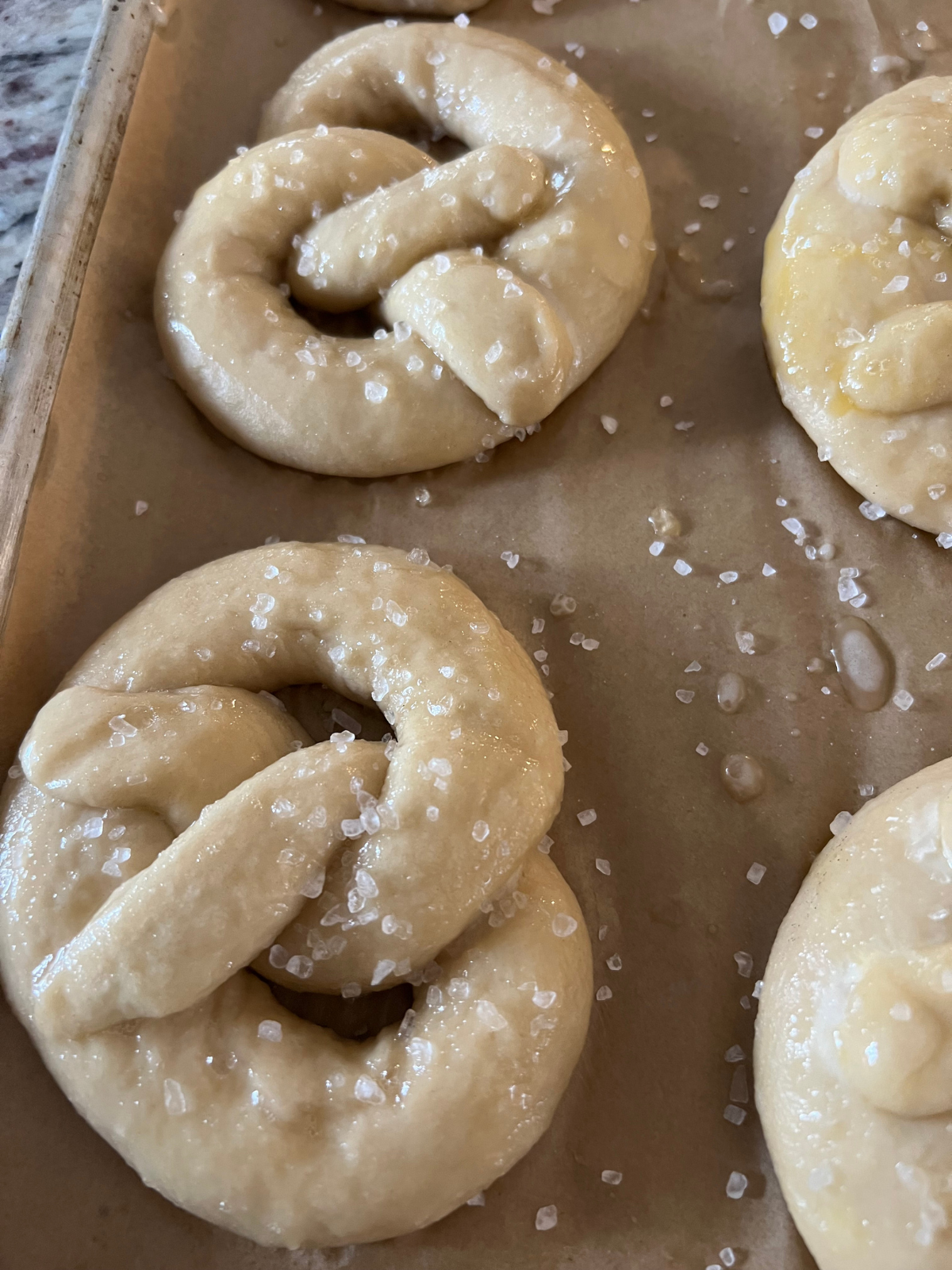 Sourdough pretzels sit unbaked on a parchment paper lined baking sheet after getting brushed with egg wash and sprinkled with coarse salt.