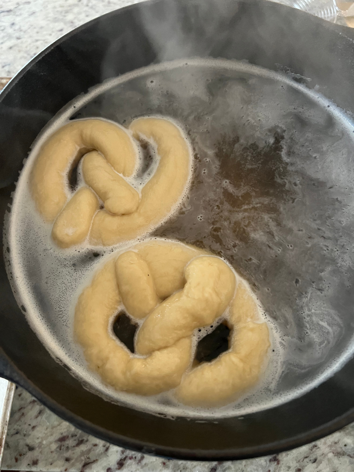 Two sourdough pretzels boil in a baking soda bath preparing to be baked.