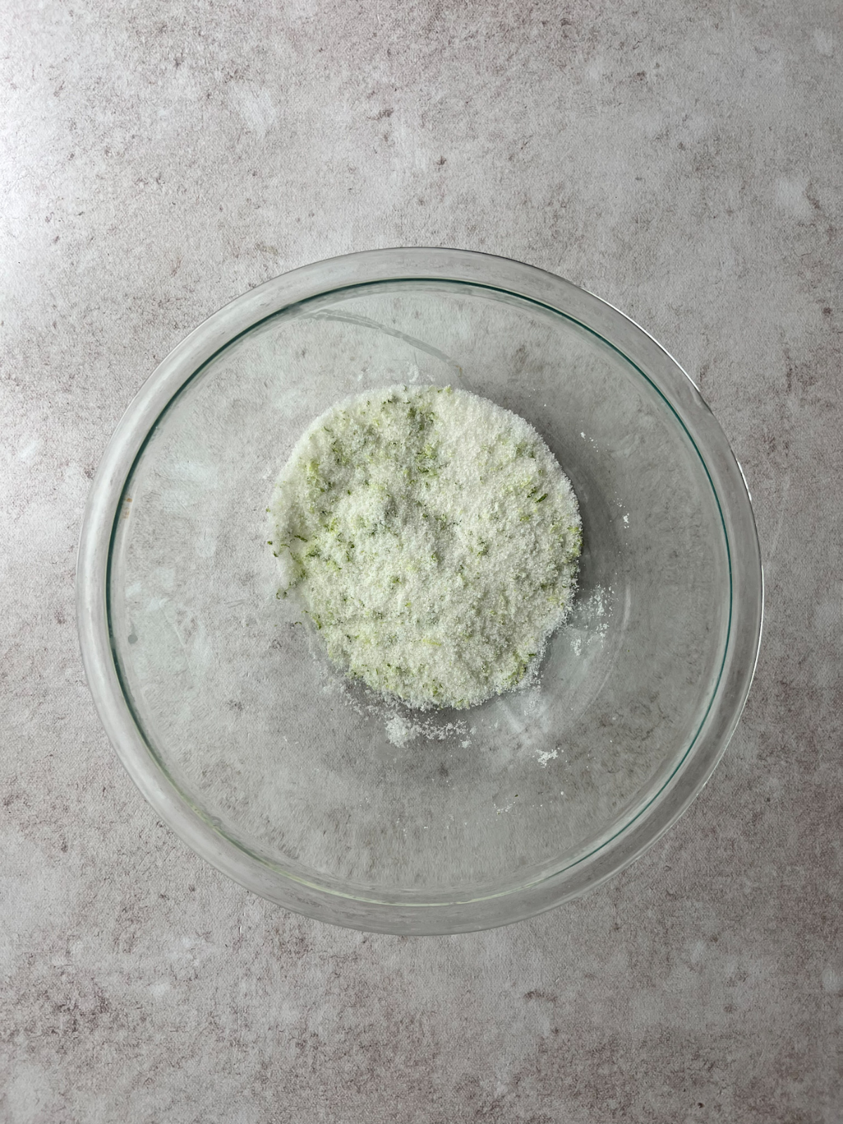 A glass mixing bowl with granulated sugar and lime zest in it.