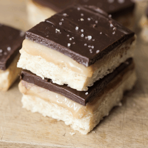 Two sourdough millionaire's shortbread bars stacked one on the other. They sit on a wooden cutting board with other bars in the background.