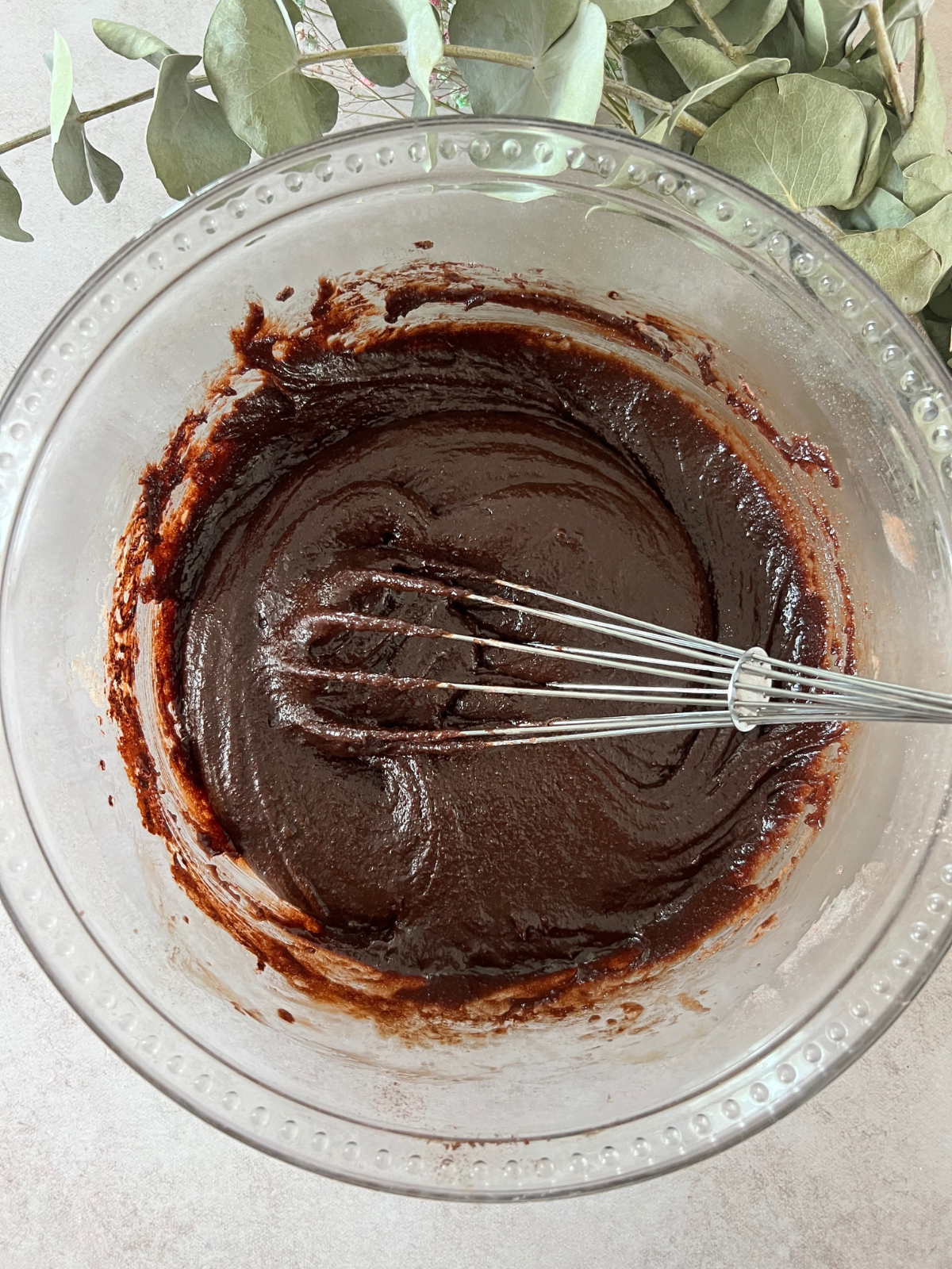 The brownie batter sits in a glass mixing bowl with a whisk in it. Greenery sits in the background.