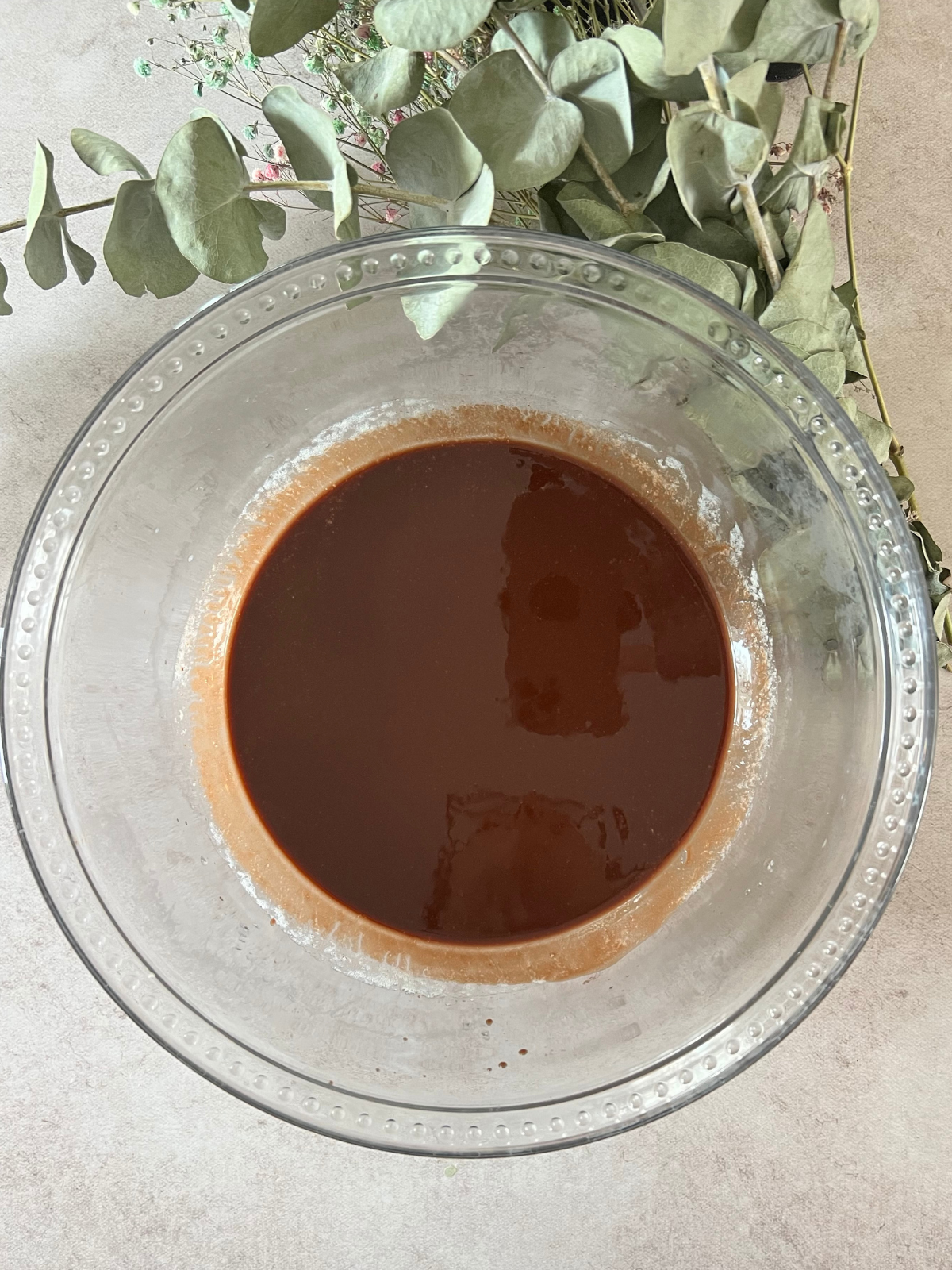 A glass mixing bowl chocolate sauce consisting of semi-sweet chocolate chips and melted butter. Greenery sits in the background.