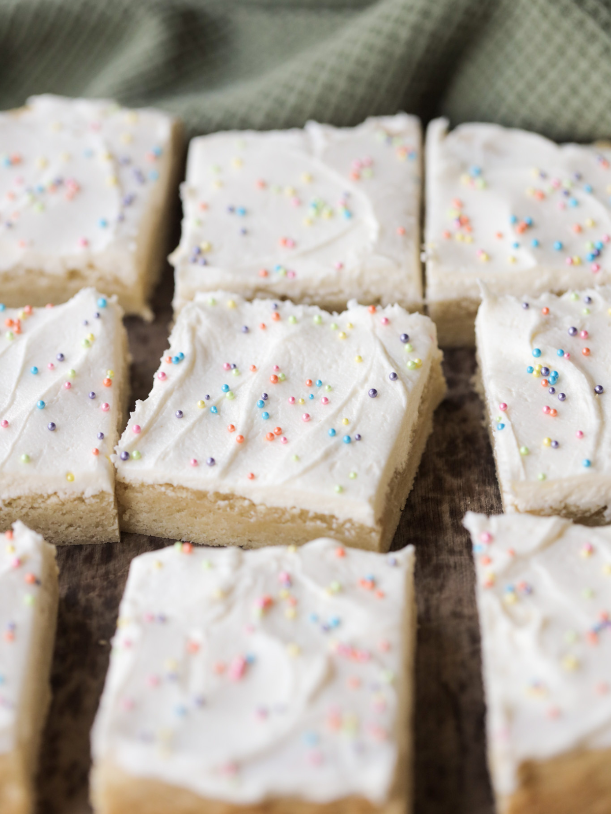 Several sourdough sugar cookie bars frosted with sprinkles sit on a wooden cutting board.