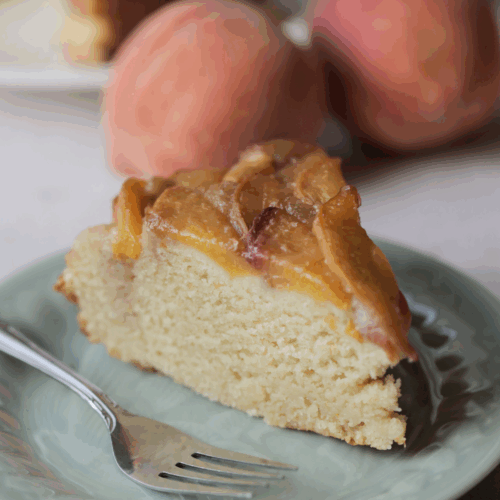 A slice of peach upside down ricotta cake on a plate with a fork. There are two peaches and the full cake in the background.