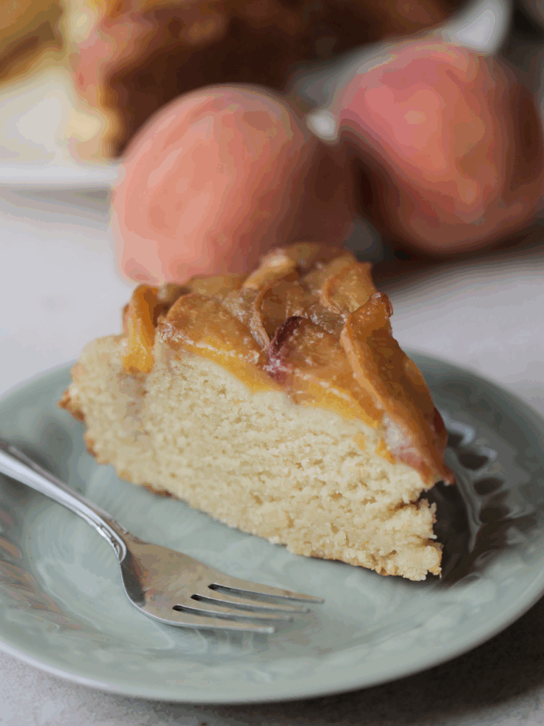 A slice of peach upside down ricotta cake on a plate with a fork. There are two peaches and the full cake in the background.