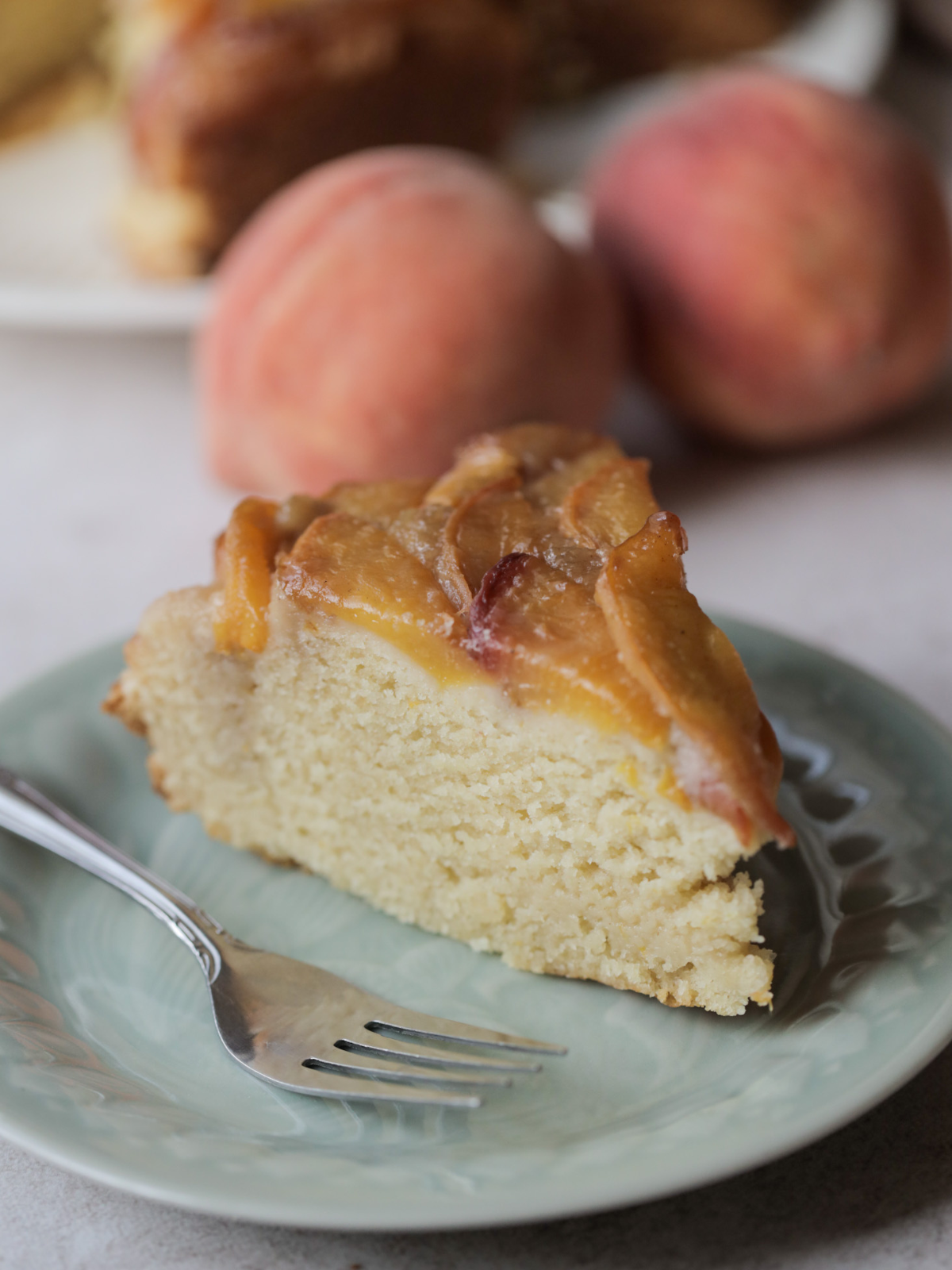 A slice of peach upside down ricotta cake on a plate with a fork. There are two peaches and the full cake in the background.