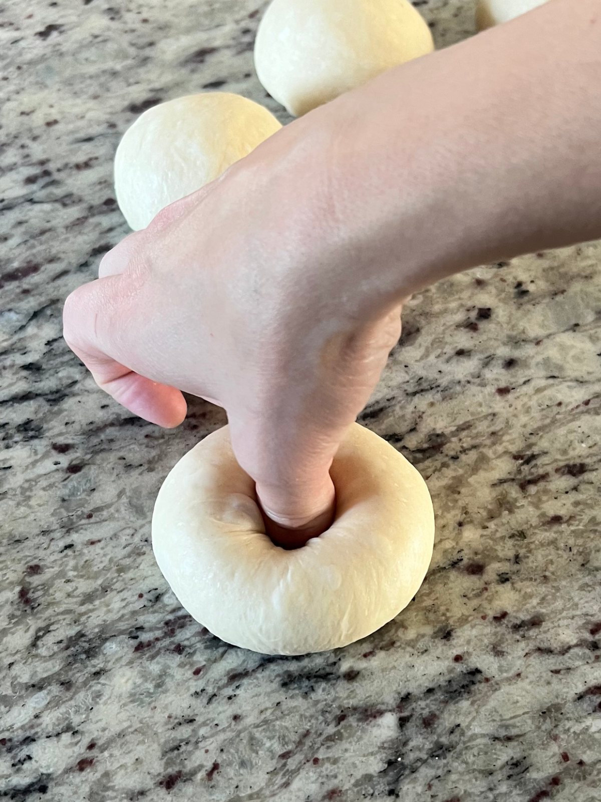 A sourdough bagel dough ball is being shaped using a thumb to poke a hole in the center of it.