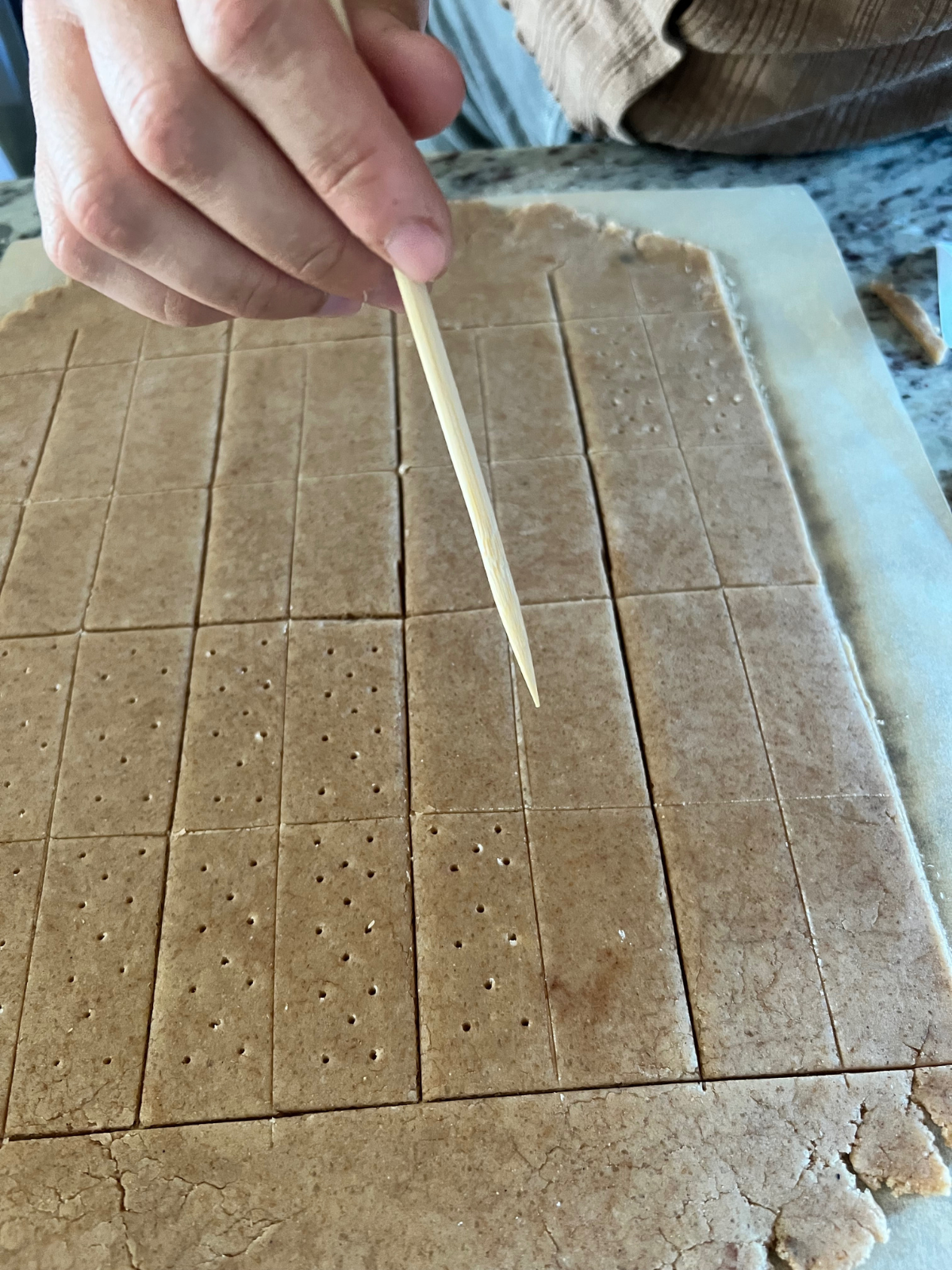 The graham cracker dough is rolled out, cut, and a hand is shown poking holes with a skewer to create the classic graham cracker look.