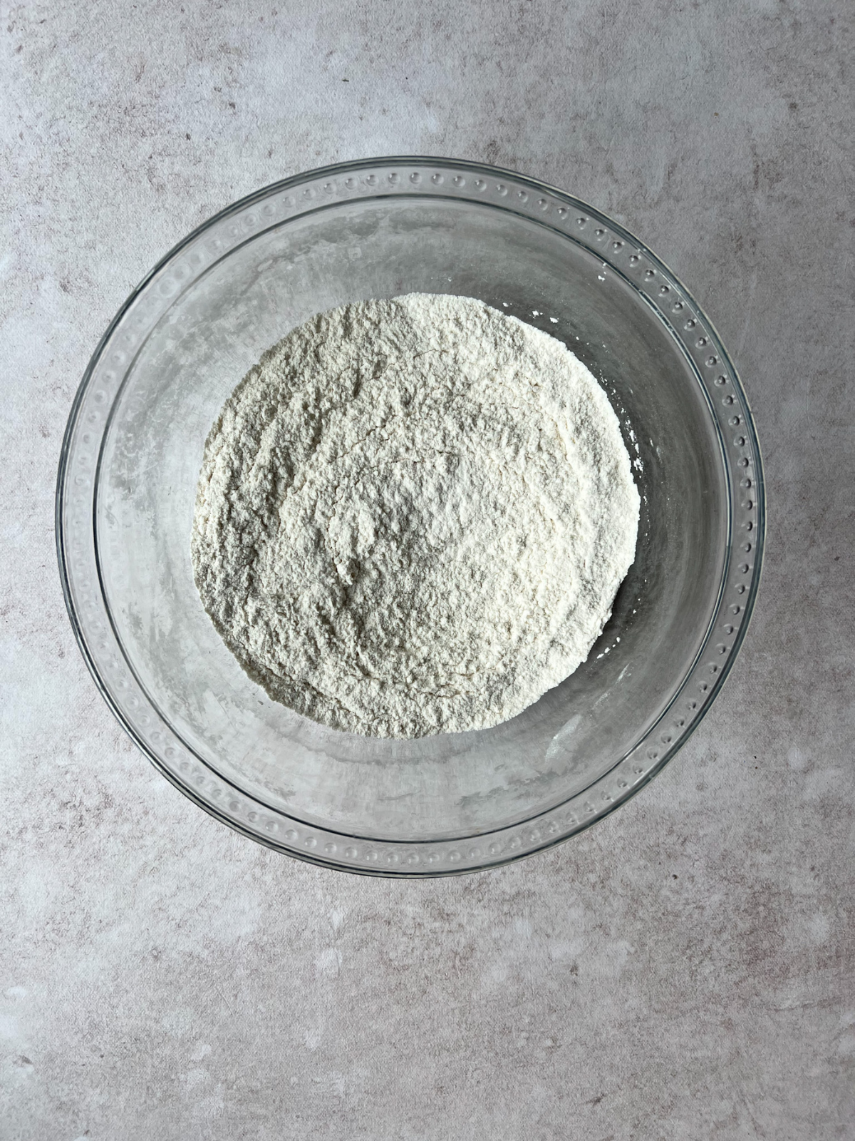 Pictured is a glass mixing bowl with the dry ingredients including flour, salt, baking powder, baking soda.