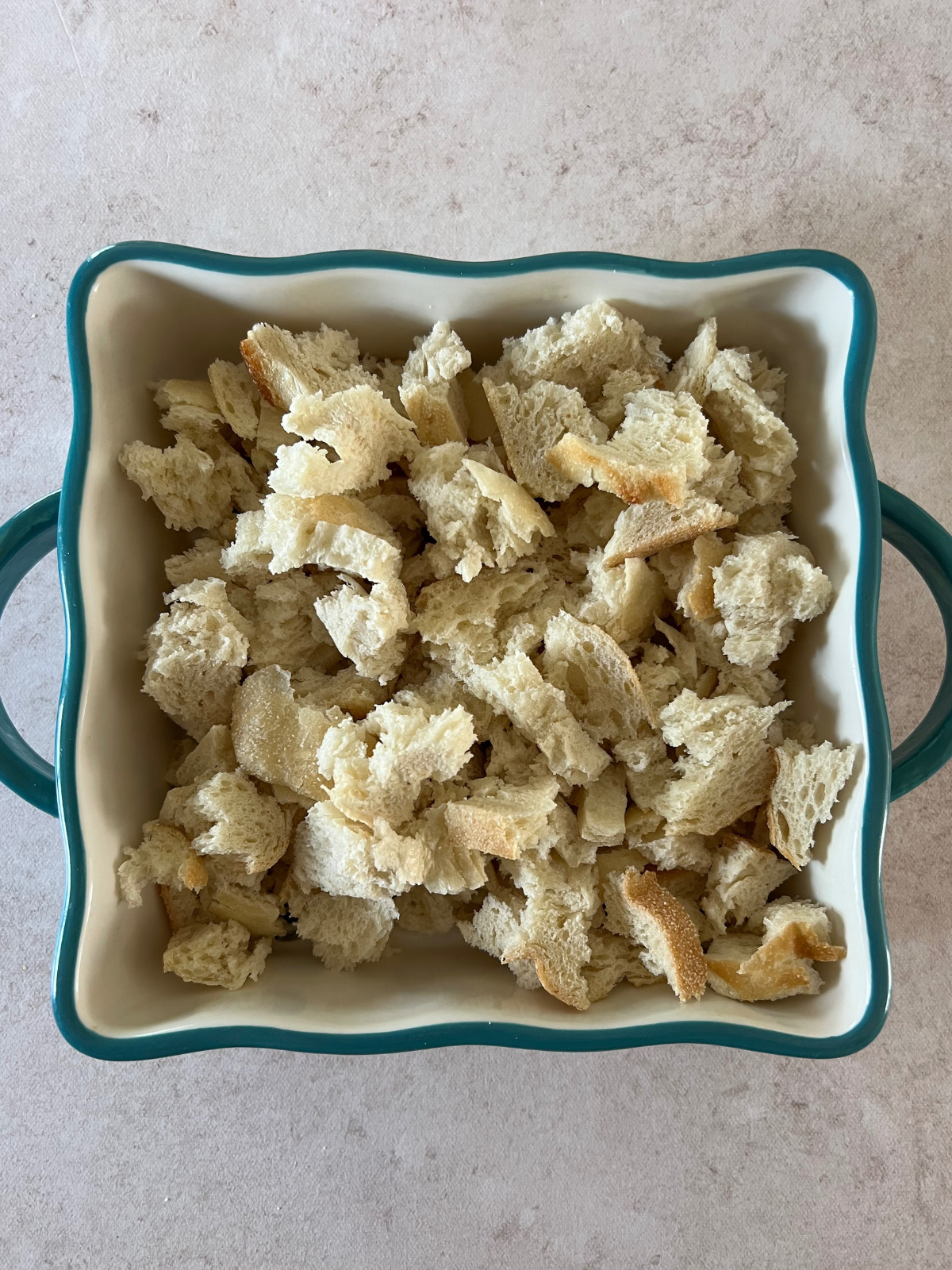 An 8x8 baking pan sits on a counter with torn apart bread pieces for the sourdough bananas foster bread pudding. 