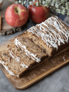 A loaf of the browned butter apple streusel loaf with three slices cut out. The loaf sits on a cutting board and apples sit in the background.