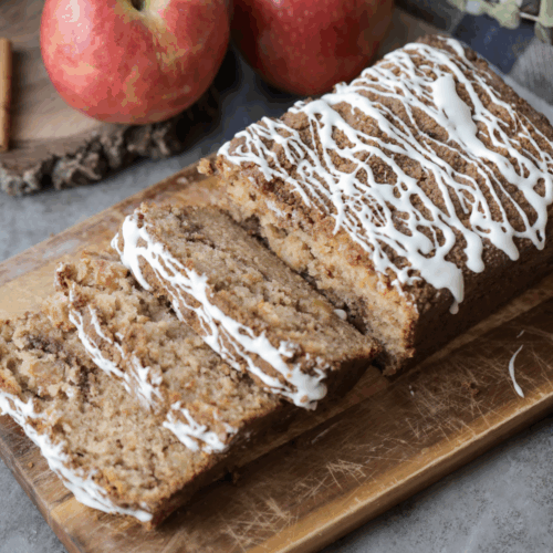 A loaf of the browned butter apple streusel loaf with three slices cut out. The loaf sits on a cutting board and apples sit in the background.