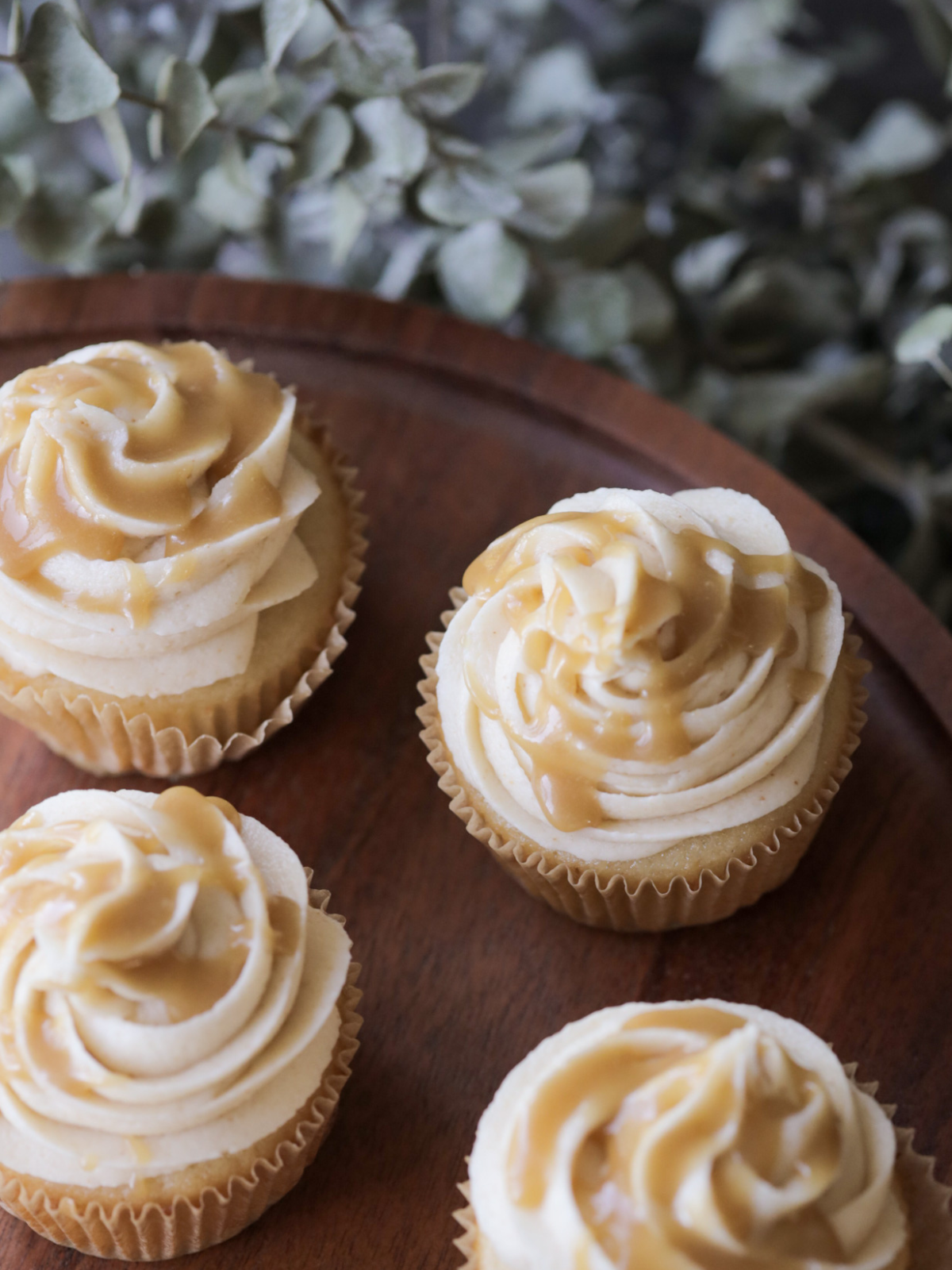 Several brown butter salted caramel cupcakes sit on a cake stand. There is caramel drizzled over the top of the cream cheese frosting.