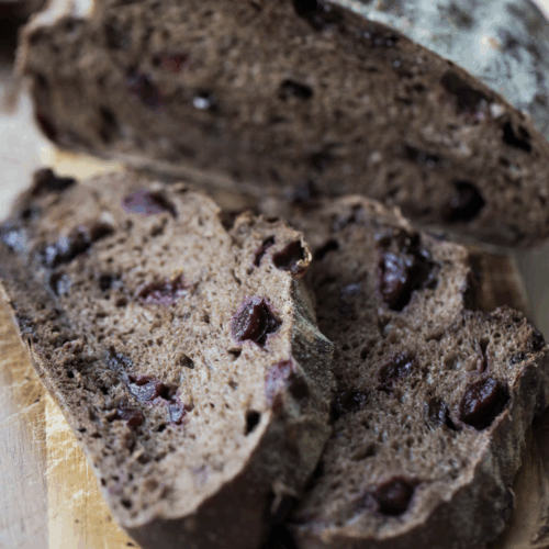 A loaf of cherry chocolate sourdough bread is cut in half in the background while two slices of the bread sit in the foreground.
