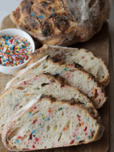 Four slices of funfetti and white chocolate sourdough bread with a bowl of sprinkles and the other half of the loaf in the background.
