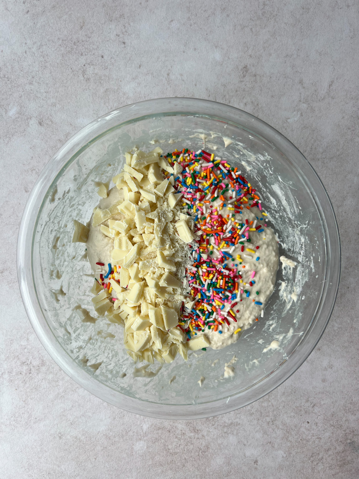 A glass mixing bowl sits on the counter with chunks of white chocolate and rainbow sprinkles in it.