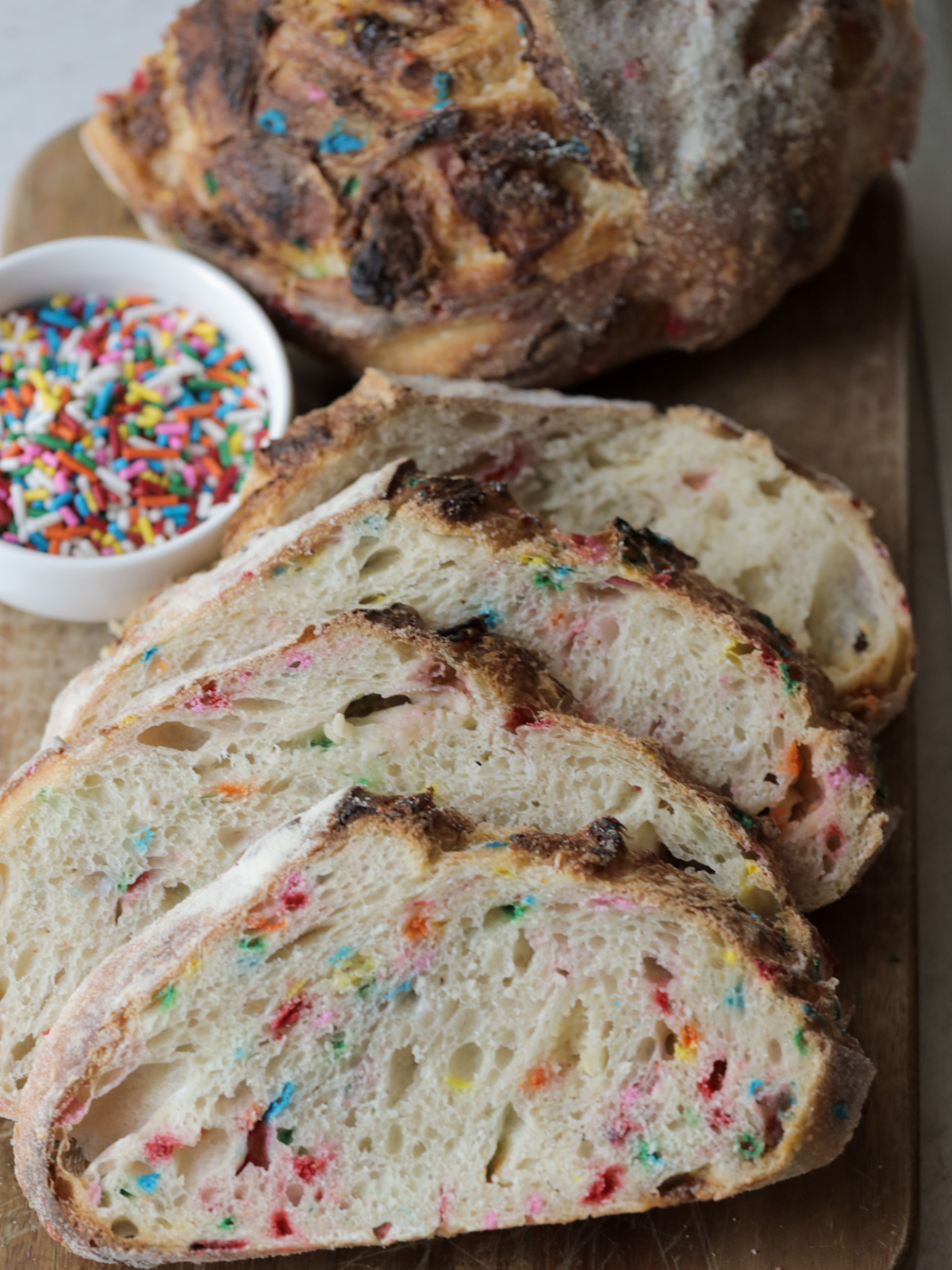 Four slices of funfetti and white chocolate sourdough bread with a bowl of sprinkles and the other half of the loaf in the background.
